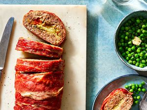 Prosciutto-Wrapped Stuffed Meatloaf on a cutting board with a knife and peas in a bowl 